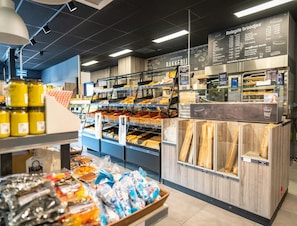 Uma área de padaria com diversos produtos de panificação expostos em prateleiras e cestas, um quadro de menu acima e uma máquina de café ao fundo.