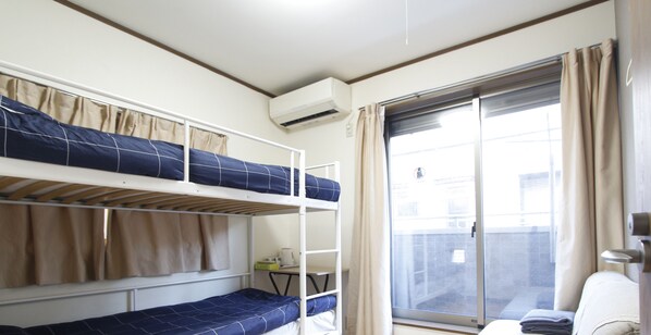 A dormitory-style room with bunk beds, a small table with a kettle, and a window with curtains.