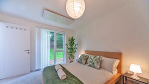 Un dormitorio con cama, mesita de noche, lámpara, ventana con persianas, puerta y planta.