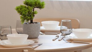Una mesa de comedor dispuesta con platos, fuentes y cubiertos blancos, una planta en maceta y una ventana con persianas.