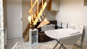 A compact living space with a wooden staircase, a small white shelf, a brown sofa, and a white ironing board.