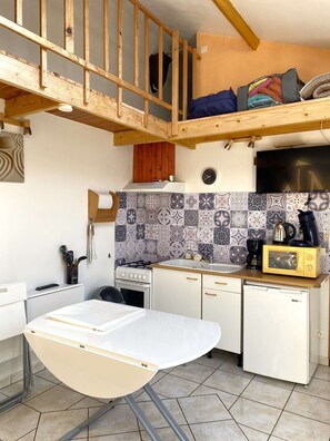 A compact kitchen with a stove, sink, and microwave. There's a wooden loft above the kitchen, and a dining table with chairs.