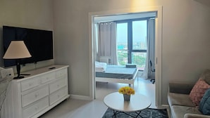 A modern living room with a flat-screen TV, a white dresser, a round coffee table with a flower vase, and a view of a balcony with a cityscape.