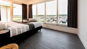 Ein Schlafzimmer mit einem großen Bett, Blick auf einen Hafen durch große Fenster und einem Balkon mit einer Tür.