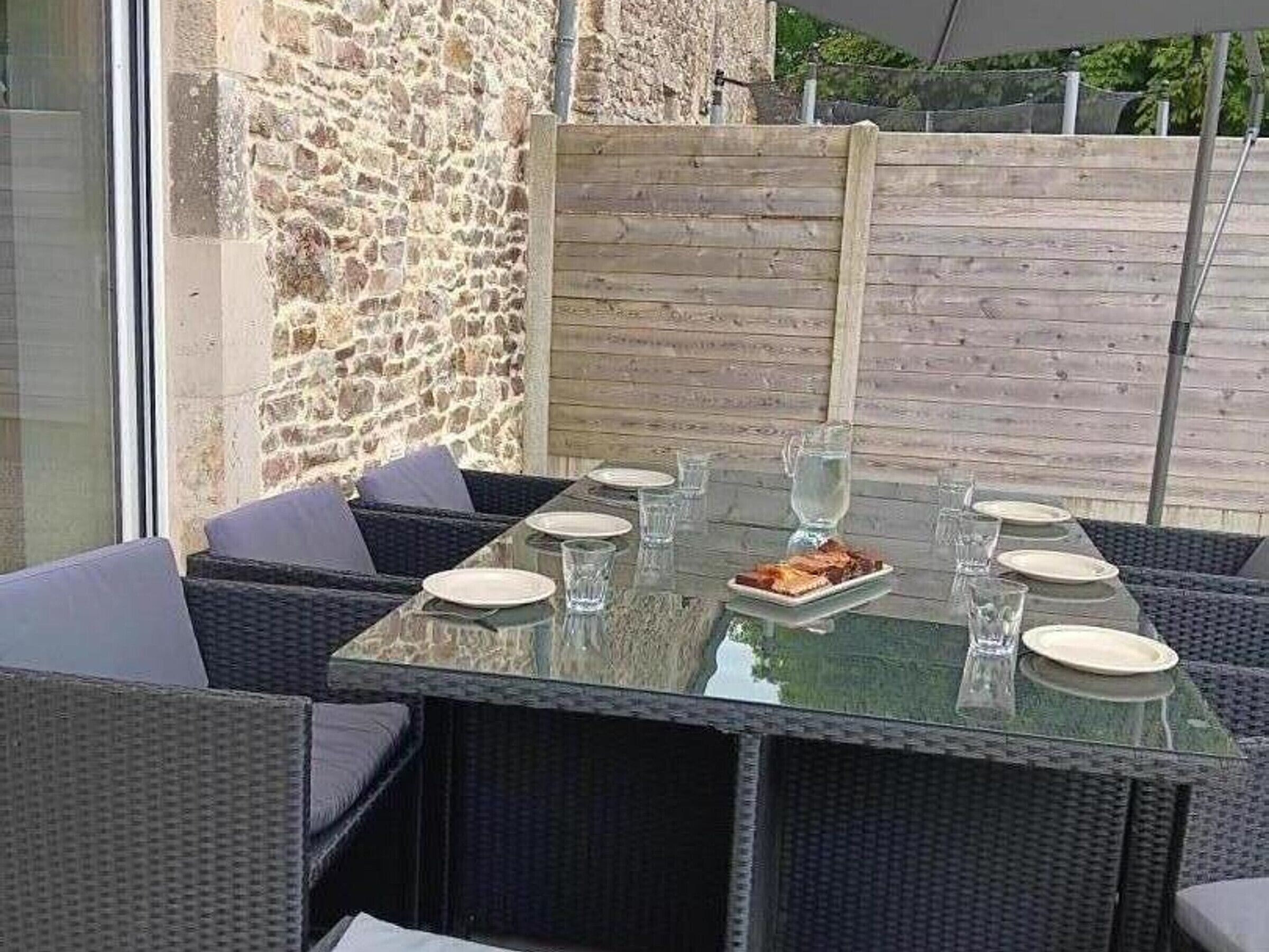 Un espace de restauration en plein air avec une table en verre et des chaises en osier, installé devant un mur de pierre et une clôture en bois.