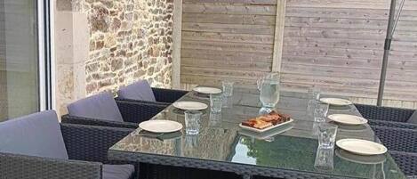 Un espace de restauration en plein air avec une table en verre et des chaises en osier, installé devant un mur de pierre et une clôture en bois.