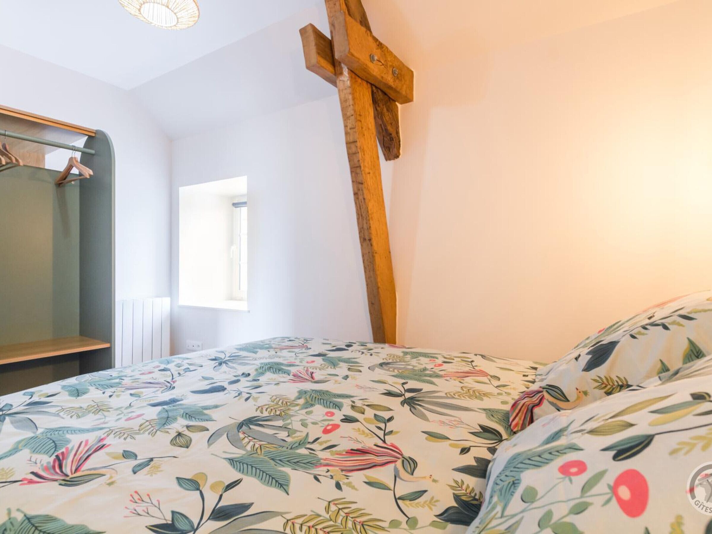 A bedroom with a floral bedspread, a wooden cross, a mirror, and a window.