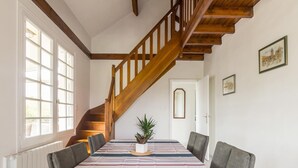 A dining room with a long table, striped tablecloth, and gray chairs. A wooden staircase leads to an upper level. - Charming house with south-facing veranda and pets allowed (Le Vast)
