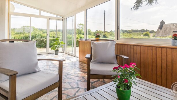 A sunlit room with large windows, wooden furniture, and a view of greenery and a building outside. - Charming house with south-facing veranda and pets allowed (Le Vast)