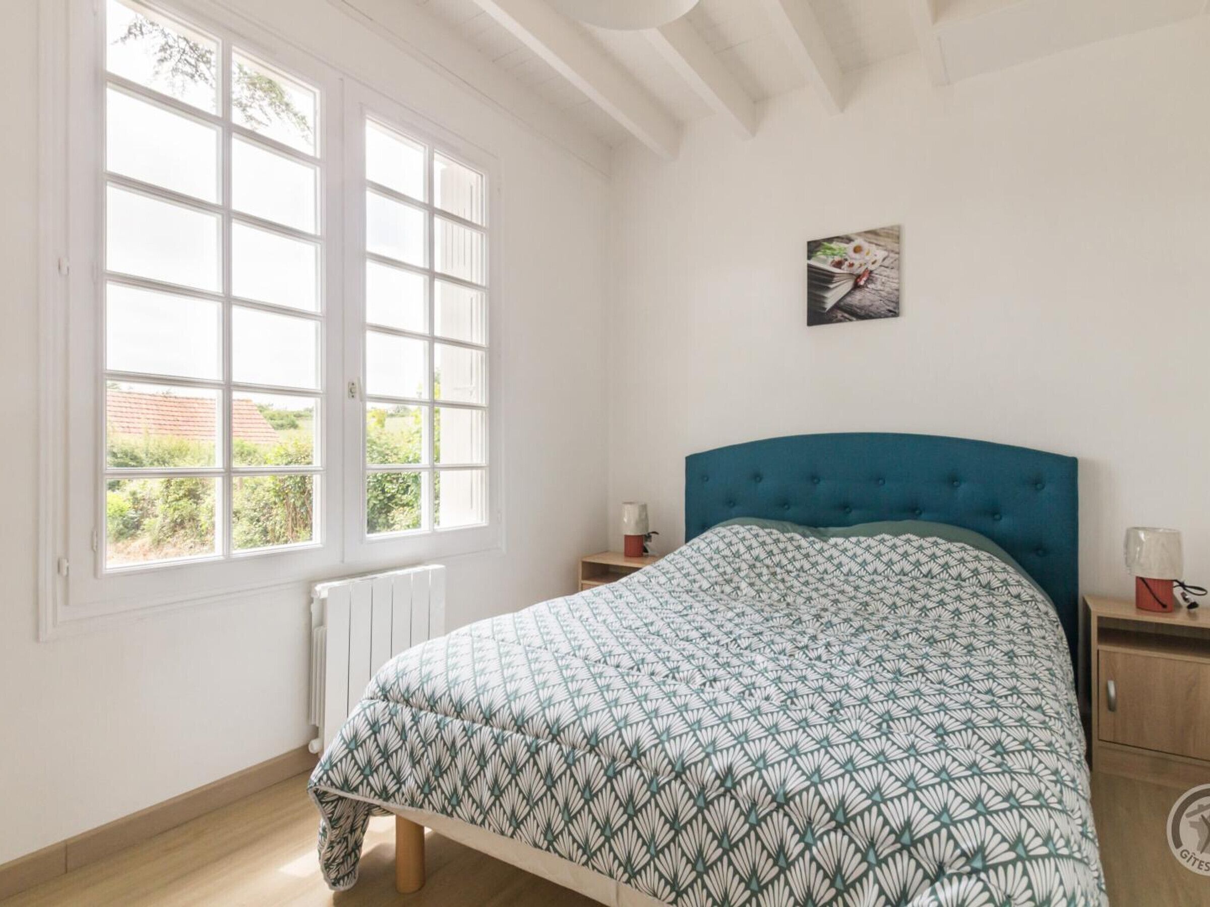 Une chambre à coucher comprenant un lit, une fenêtre, un radiateur, une table de chevet et un tableau accroché au mur.