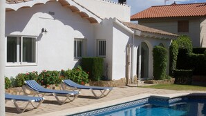 Un espace aménagé au bord de la piscine, avec des chaises longues, une piscine et un bâtiment blanc doté d’un toit en tuiles.