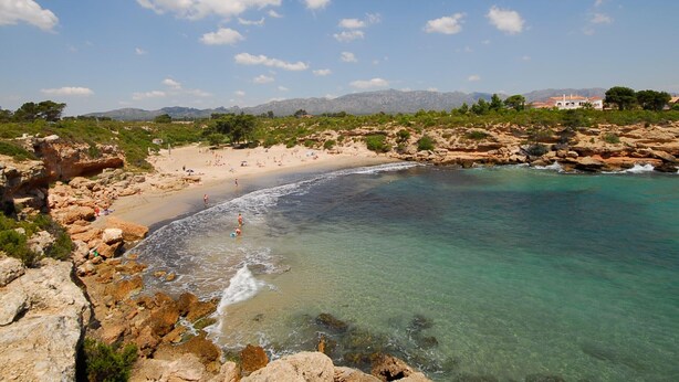Une plage côtière au paysage rocheux, avec une eau turquoise et limpide, et des montagnes qui se dessinent à l’horizon.
