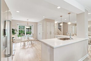 A modern kitchen with a central island, a dining area with chairs, and a view of greenery outside.