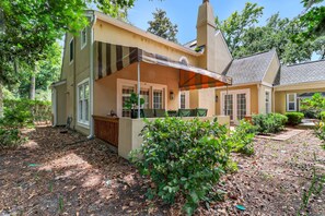 Ein zweistöckiges Haus mit überdachter Terrasse, Schornstein und Garten mit verschiedenen Pflanzen.