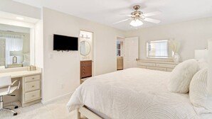 Un dormitorio con cama, escritorio, silla, televisor, ventilador de techo y ventana.