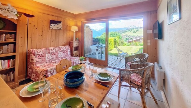 Un comedor acogedor con una mesa de madera dispuesta para dos personas, un sofá y una estantería. Un amplio ventanal ofrece una vista panorámica de un paisaje montañoso.