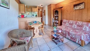 Una sala de estar acogedora con un sofá, una mesa de comedor para cuatro, una estantería y una cocina al fondo.