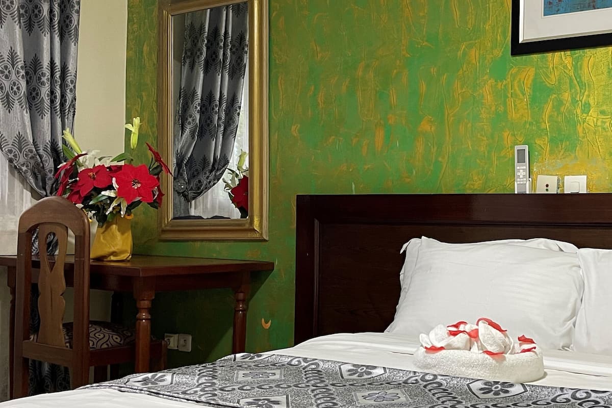 A hotel room with a bed, a nightstand, a chair, a vase with flowers, and a painting on the wall.