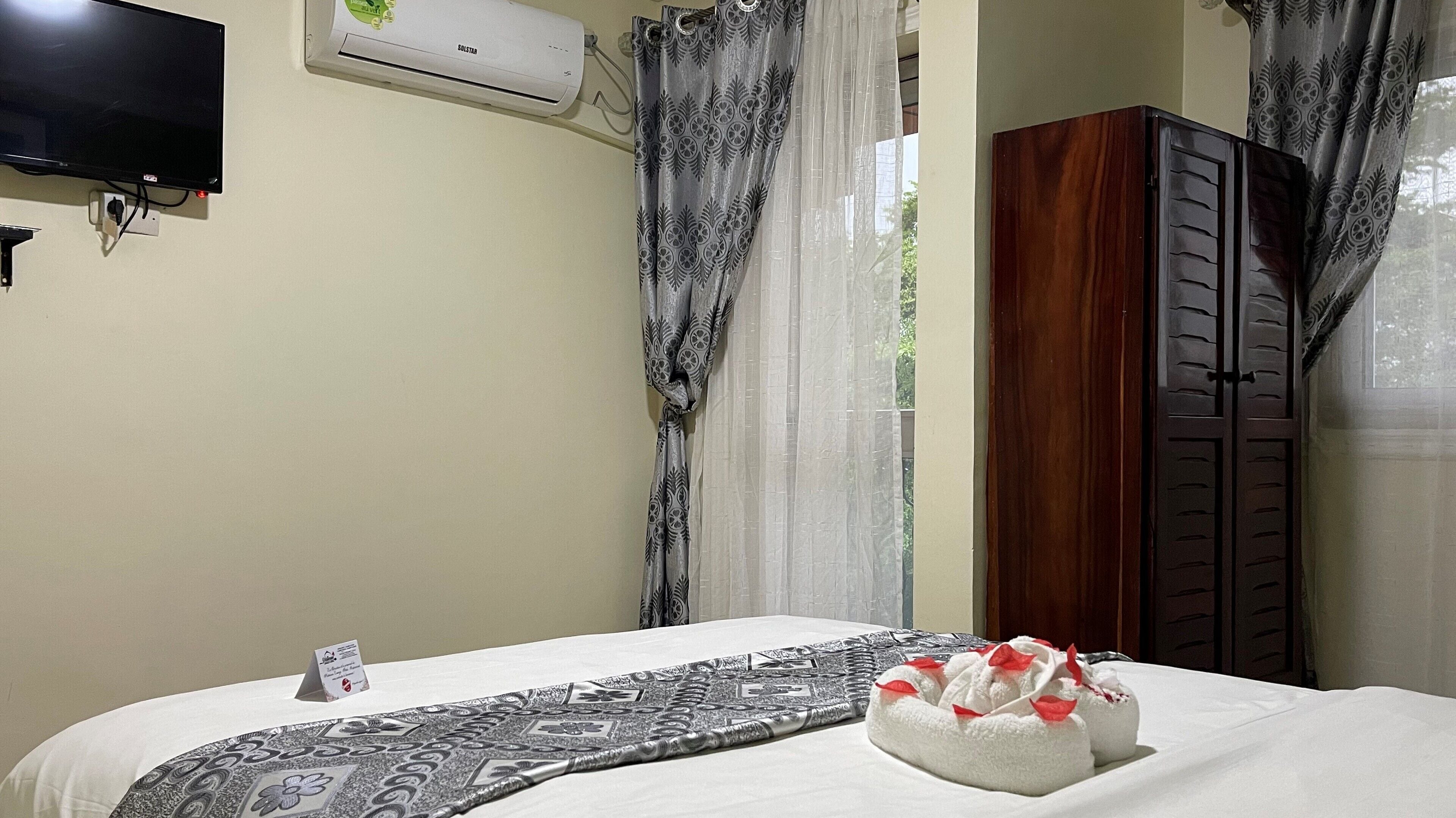 A hotel room with a bed, a TV mounted on the wall, an air conditioner, a wooden wardrobe, and a window with curtains.