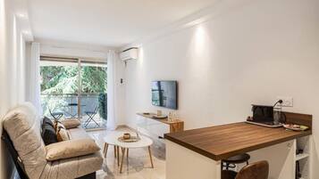 A modern living room with a sofa, a coffee table, and a TV. There is a balcony with outdoor furniture.