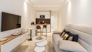 A modern living room with a sofa, a TV mounted on the wall, and a kitchen area in the background. - Le Center avec joli balcon et parking (Cannes)