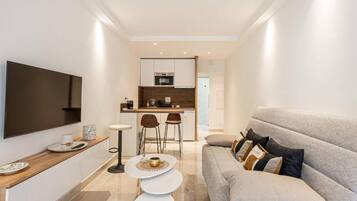A modern living room with a sofa, a TV mounted on the wall, and a kitchen area in the background.