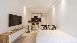 A modern hotel room with a bed, a TV mounted on the wall, a small kitchen area, and a seating space with chairs. - Le Center avec joli balcon et parking (Cannes)