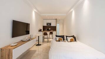 A modern hotel room with a bed, a TV mounted on the wall, a small kitchen area, and a seating space with chairs.