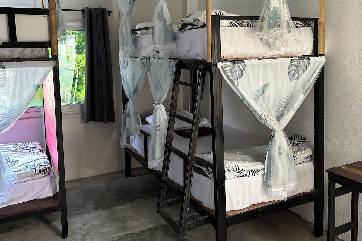 A room with bunk beds, each with a mosquito net. There is a window with curtains, a wall-mounted air conditioner, and a concrete floor.