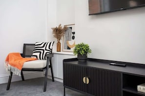 A modern living room with a sofa, a black and white patterned pillow, a potted plant, a remote control, and a framed artwork on the wall.