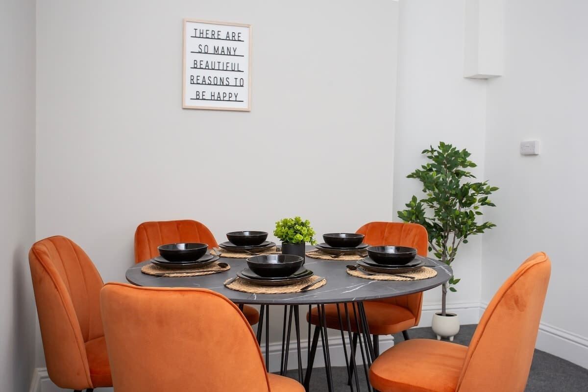 Un comedor con una mesa redonda, seis sillas anaranjadas, cuencos negros y una planta en maceta.