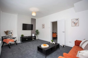 A modern living room with a flat-screen TV, a black console table, an orange sofa, and a grey carpet.
