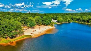 A lakeside view with a sandy beach, dense greenery, and a clear blue sky. - The Firefly – A-Frame Cabin with Hot Tub, Arcade & 3 Living Rooms (Albrightsville)