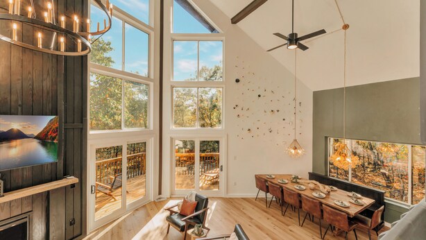 A spacious living area with a large TV, wooden flooring, a dining table, and a balcony with a view of trees. - The Firefly – A-Frame Cabin with Hot Tub, Arcade & 3 Living Rooms (Albrightsville)