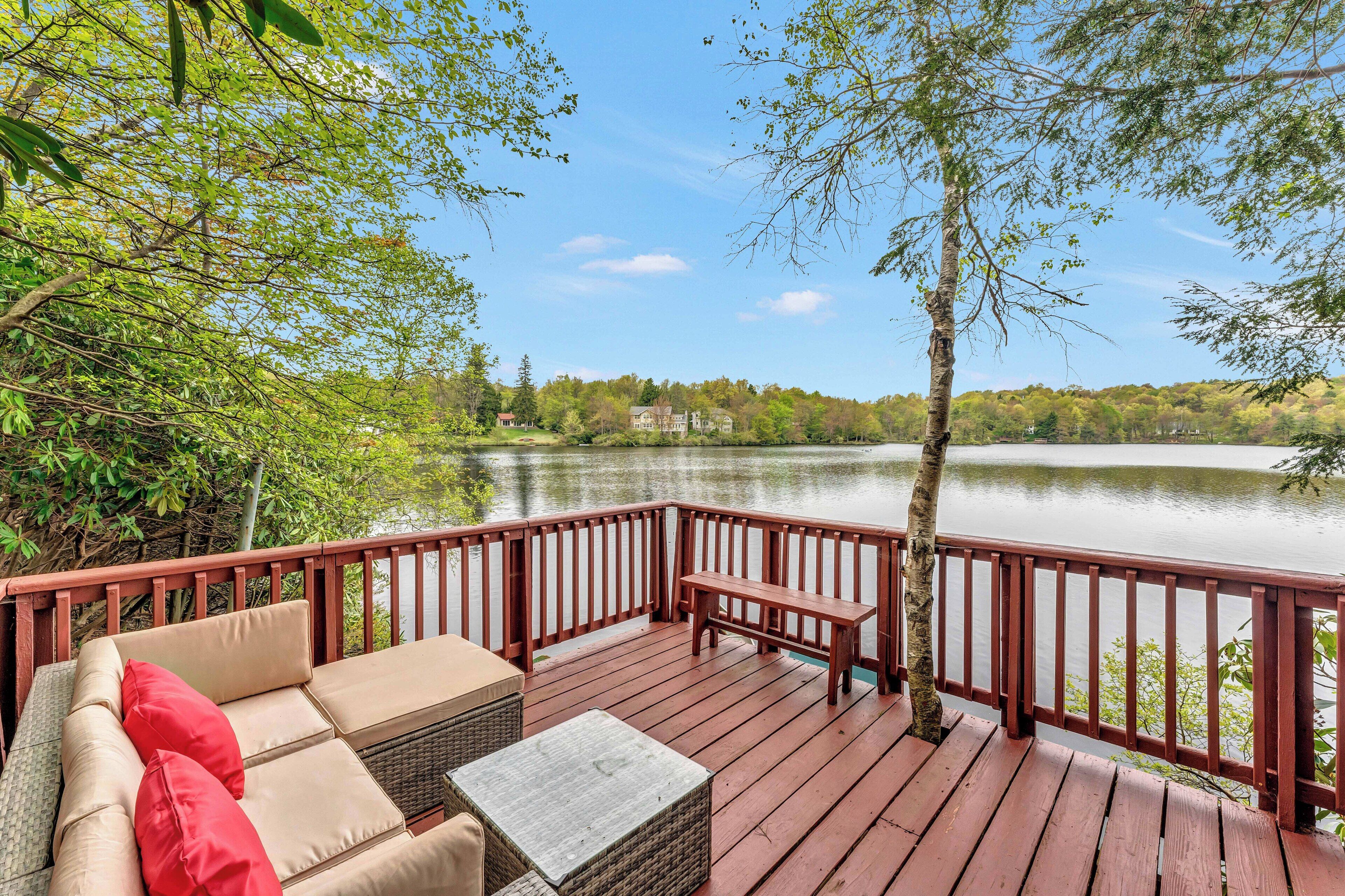 A wooden deck with a sofa and a bench, overlooking a lake with trees and a distant building.