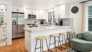 A modern kitchen with white cabinets, stainless steel appliances, a large island with stools, and a clock on the wall. - Green Lake Remodelled home with AC, garage, yard (Seattle)