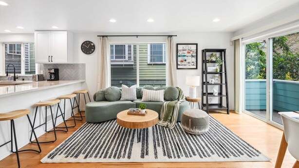A modern living room with a green sofa, a round coffee table, and a striped rug. There is a kitchen area with white cabinets and a bar stool. A bookshelf and a ladder are also visible. - Green Lake Remodelled home with AC, garage, yard (Seattle)