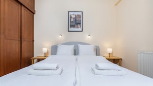 Una cama doble impecablemente arreglada con sábanas blancas, dos mesitas de noche con lámparas, un cuadro enmarcado sobre la cama y un armario de madera a la izquierda.