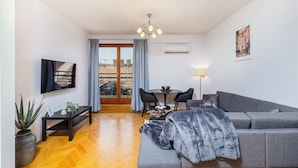 Una sala de estar moderna con un sofá gris, una mesita con sillas, un televisor montado en la pared y un balcón con vistas a edificios.