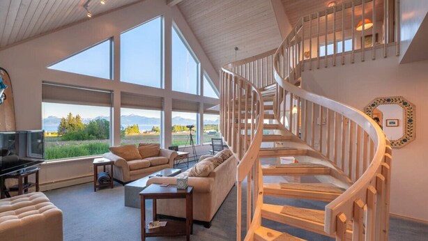 A spacious living room with a large window, a wooden staircase, and a television. - Feathers + Fireweed (Homer)