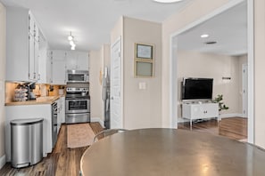A modern kitchen with stainless steel appliances, a dining area with a round table, and a flat-screen TV in a living space. - Ufer Haus - Game Room, Fire Pit, Walk to Main (Fredericksburg)