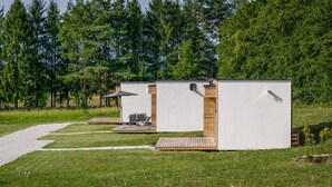 Una hilera de cabañas modernas y blancas con terrazas de madera y zonas de estar al aire libre, rodeadas por un campo de césped y árboles frondosos.