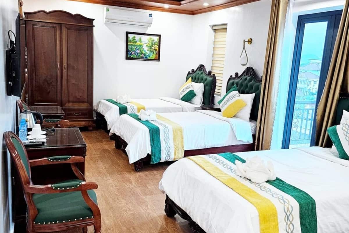 Une chambre d’hôtel avec trois lits, une armoire en bois, un bureau avec des chaises et une vue sur la ville par la fenêtre.