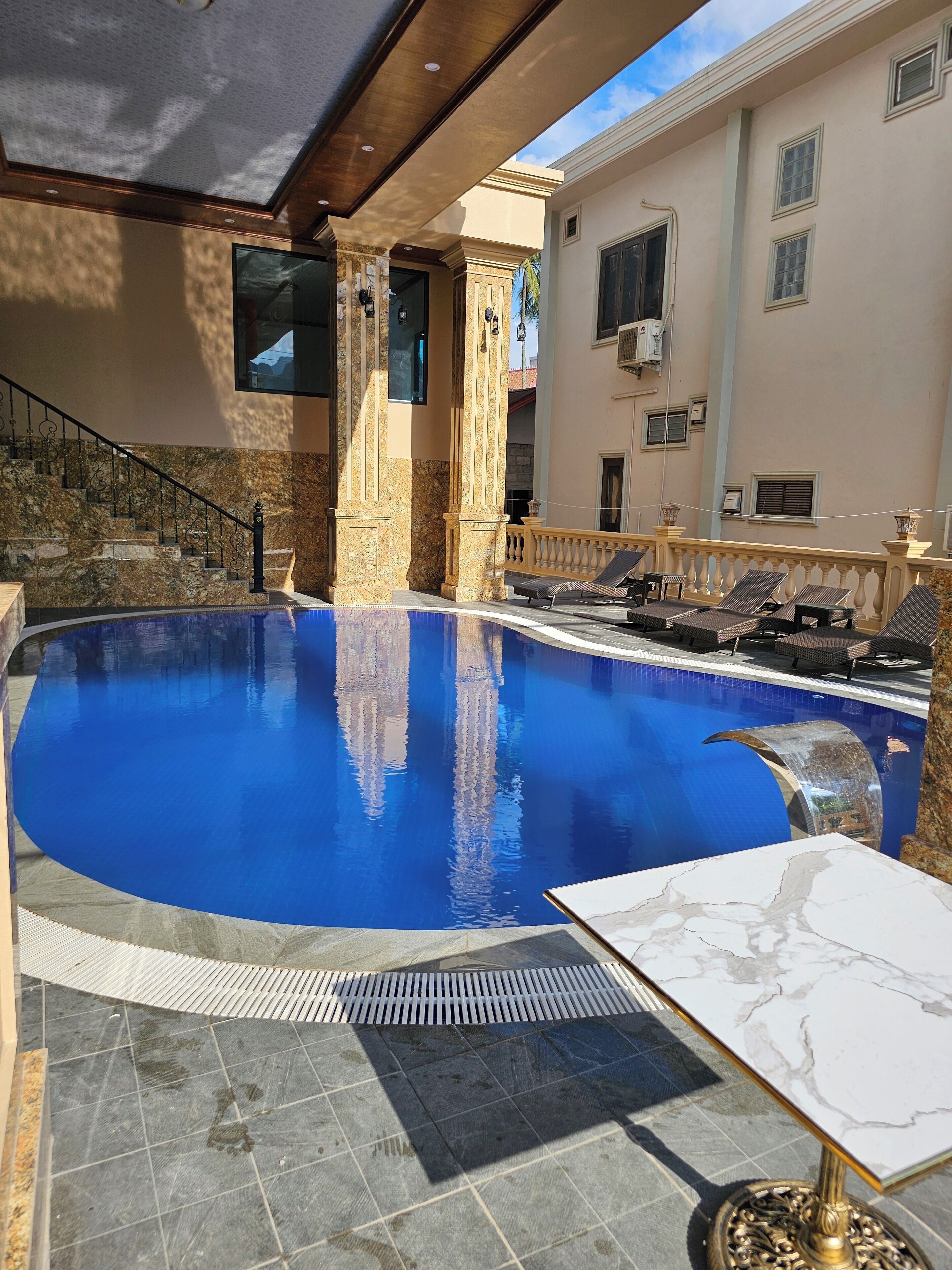 A swimming pool with a curved edge, a marble-tiled floor, and a sunlit outdoor area with lounge chairs and a table.
