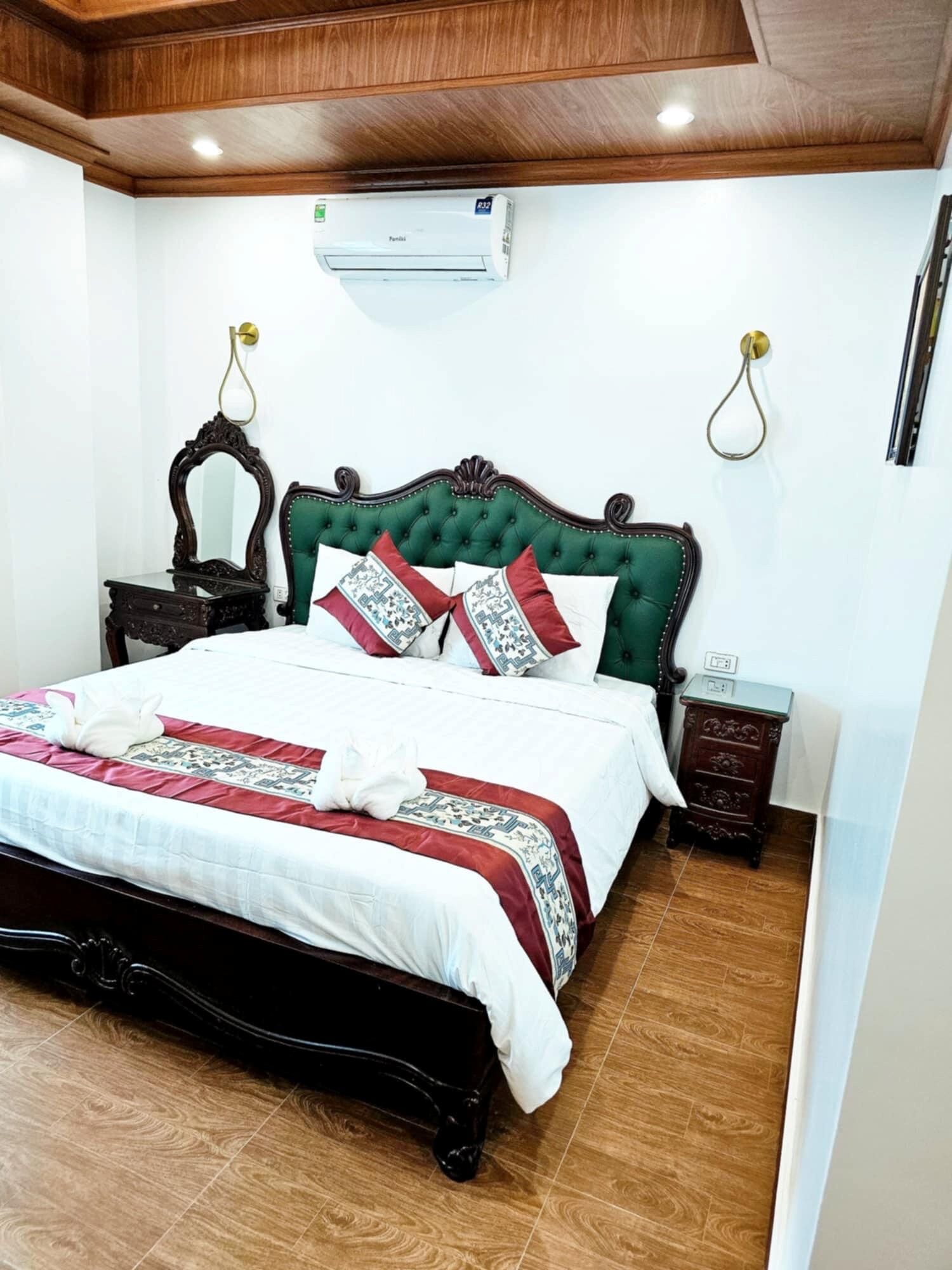 A neatly made bed with a green upholstered headboard, a white comforter, and patterned pillows. There is a wooden nightstand with a small table beside the bed, and an air conditioning unit mounted on the wall above.
