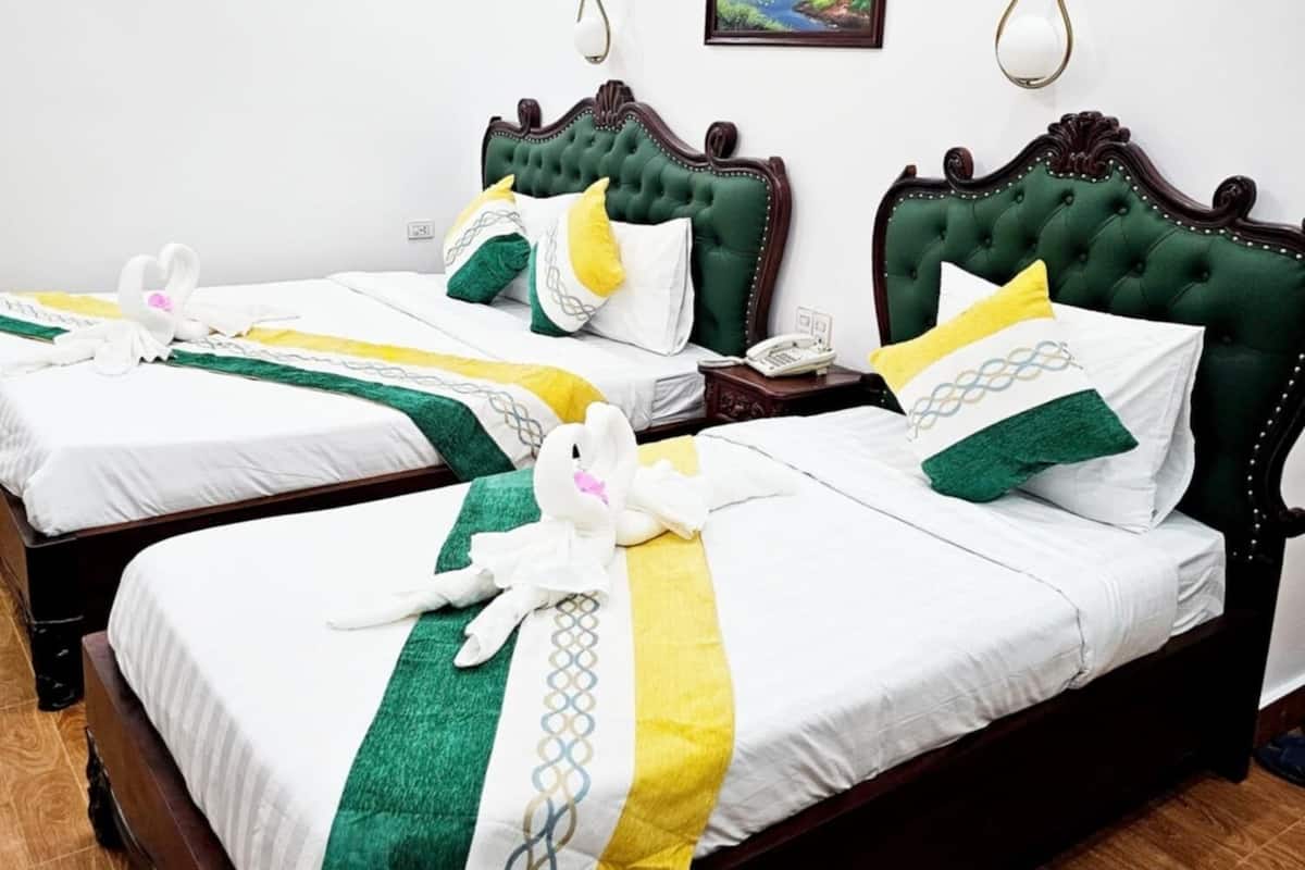 Une chambre d’hôtel avec deux lits, chacun doté d’une tête de lit verte et de draps blancs. Il y a un tableau au mur, un ventilateur de plafond et un téléphone sur la table de chevet.