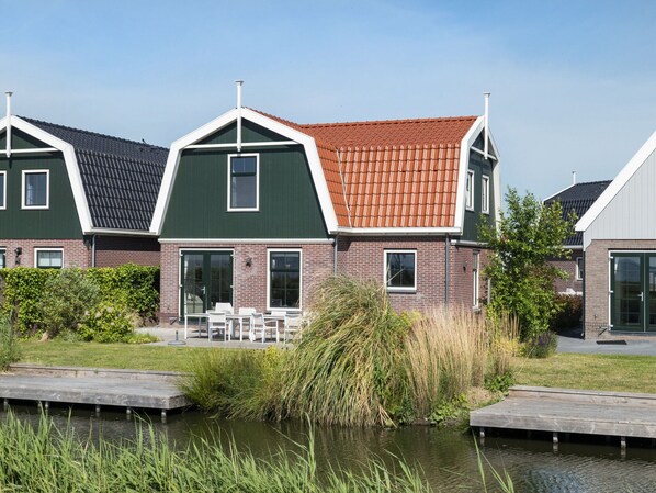 A row of modern houses with green and red roofs, a grassy area, and a water body in the foreground. - Holiday Home With Sauna Near Amsterdam (Uitdam)