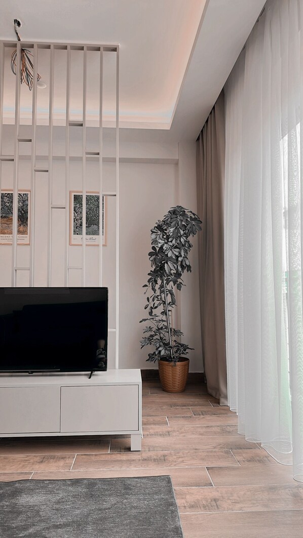 A modern living room with a flat-screen TV, a potted plant, and a large window with sheer curtains. - Vista Living Apartments (Trabzon)