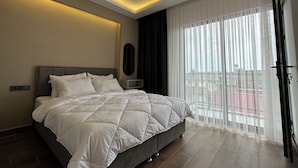 A modern hotel room with a large bed, a ceiling light, a window with sheer curtains, and a view of the outside balcony. - Vista Living Apartments (Trabzon)