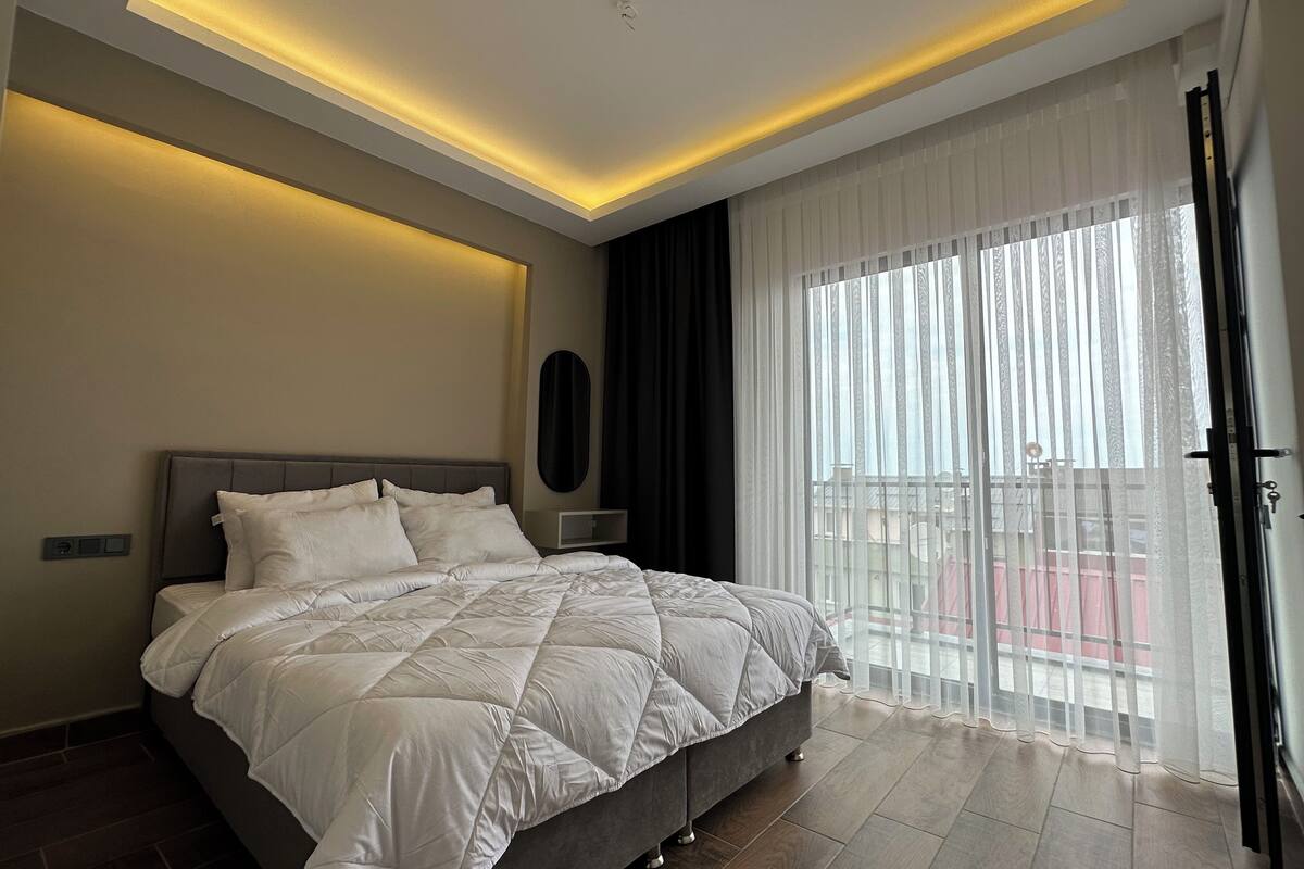 A modern hotel room with a large bed, a ceiling light, a window with sheer curtains, and a view of the outside balcony.